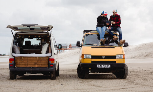 Zwei VW-Bullis stehen am Strand, auf einem sitzen zwei Personen