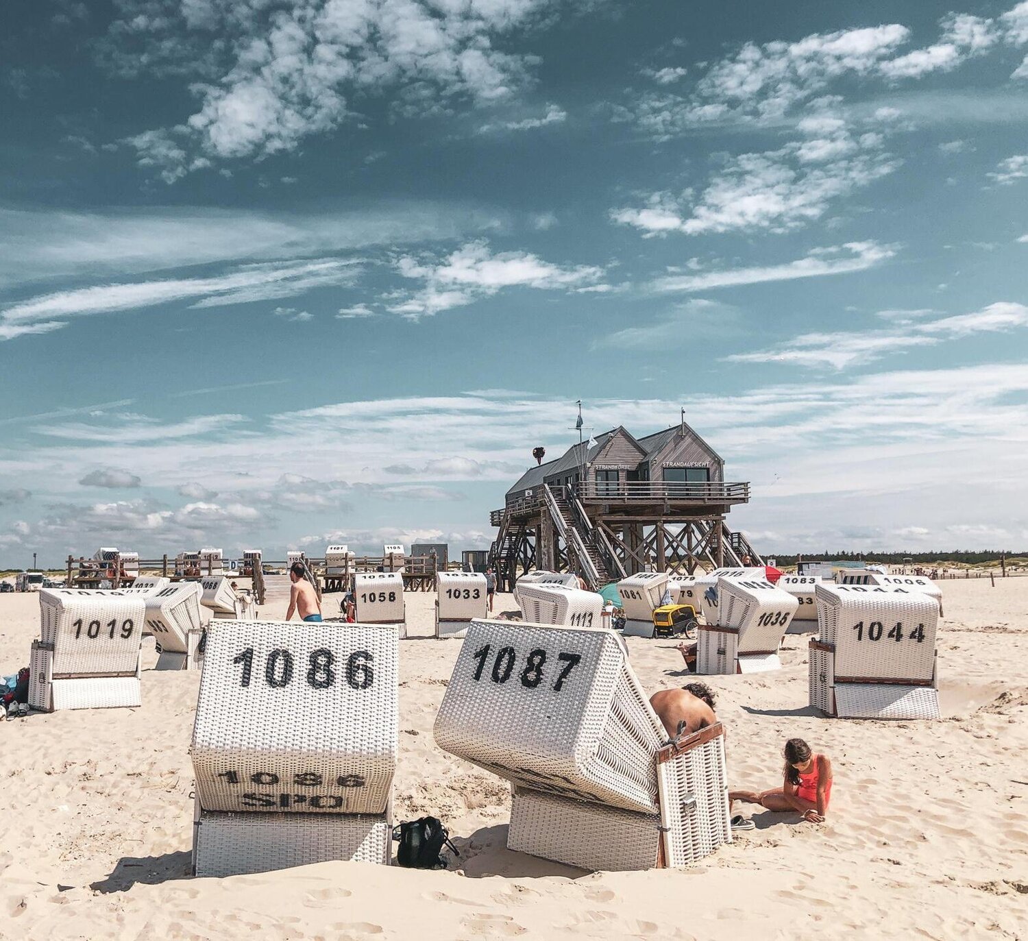 Ein sonniger Tag am Strand. In den Strandkörben genießen die Strandbesucher die Sonnenstrahlen auf der Haut. Im Hintergrund steht ein kultiger Pfahlbau.
