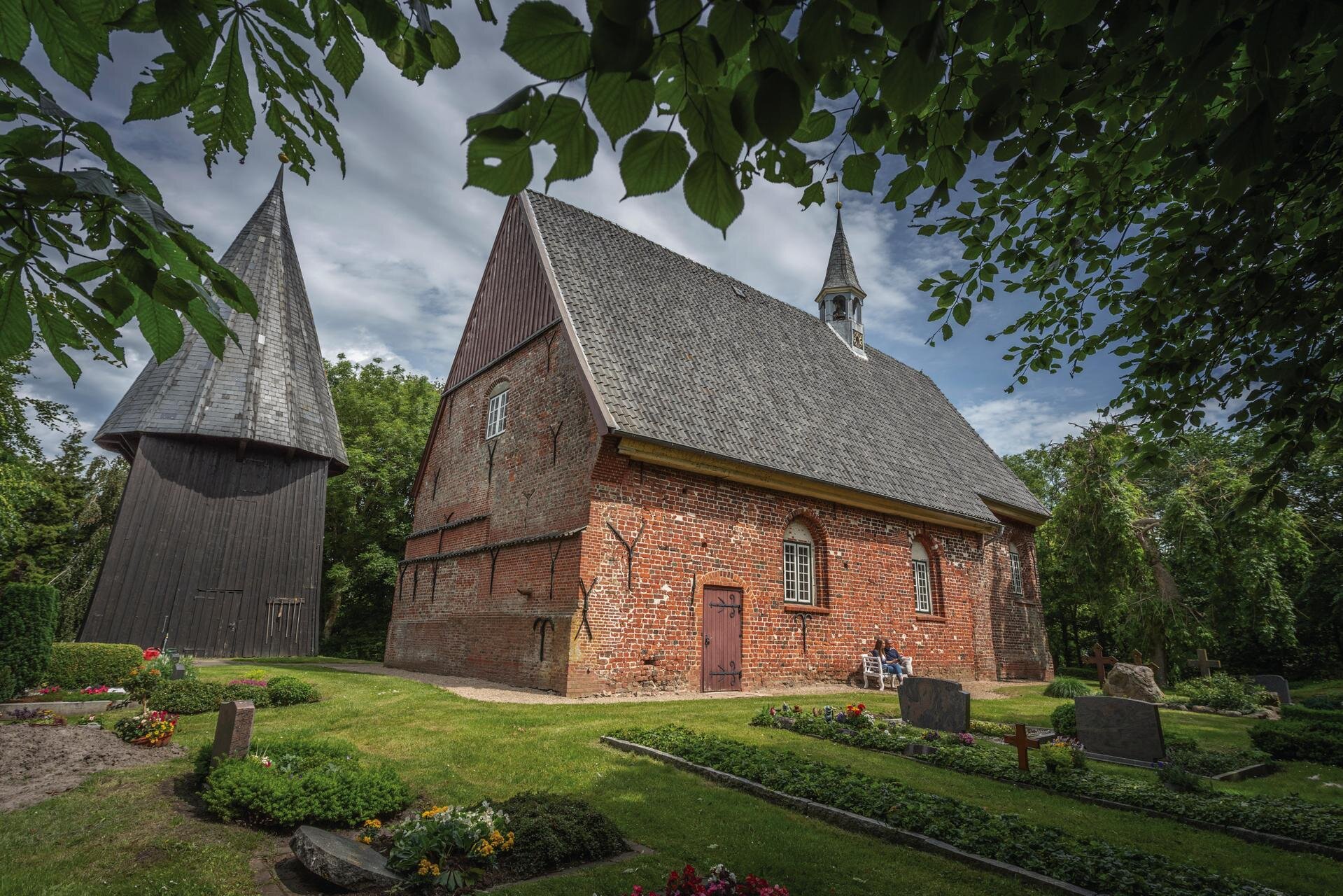 urige Kirche aus rotem Backstein mit dunklem Dach und kleiner Kirchturmspitze und separatem Turm daneben