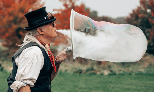 Ein Mann mit Hut bläst Rauch in eine sehr große Seifenblase.