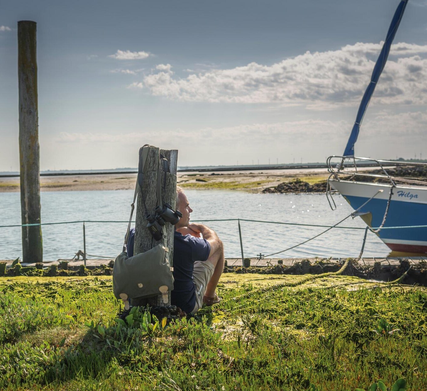 Mann sitzt an einen kleinen Pfahl gelehnt im Gras und blickt auf das Wasser im Hafen vom Tetenbüllspieker