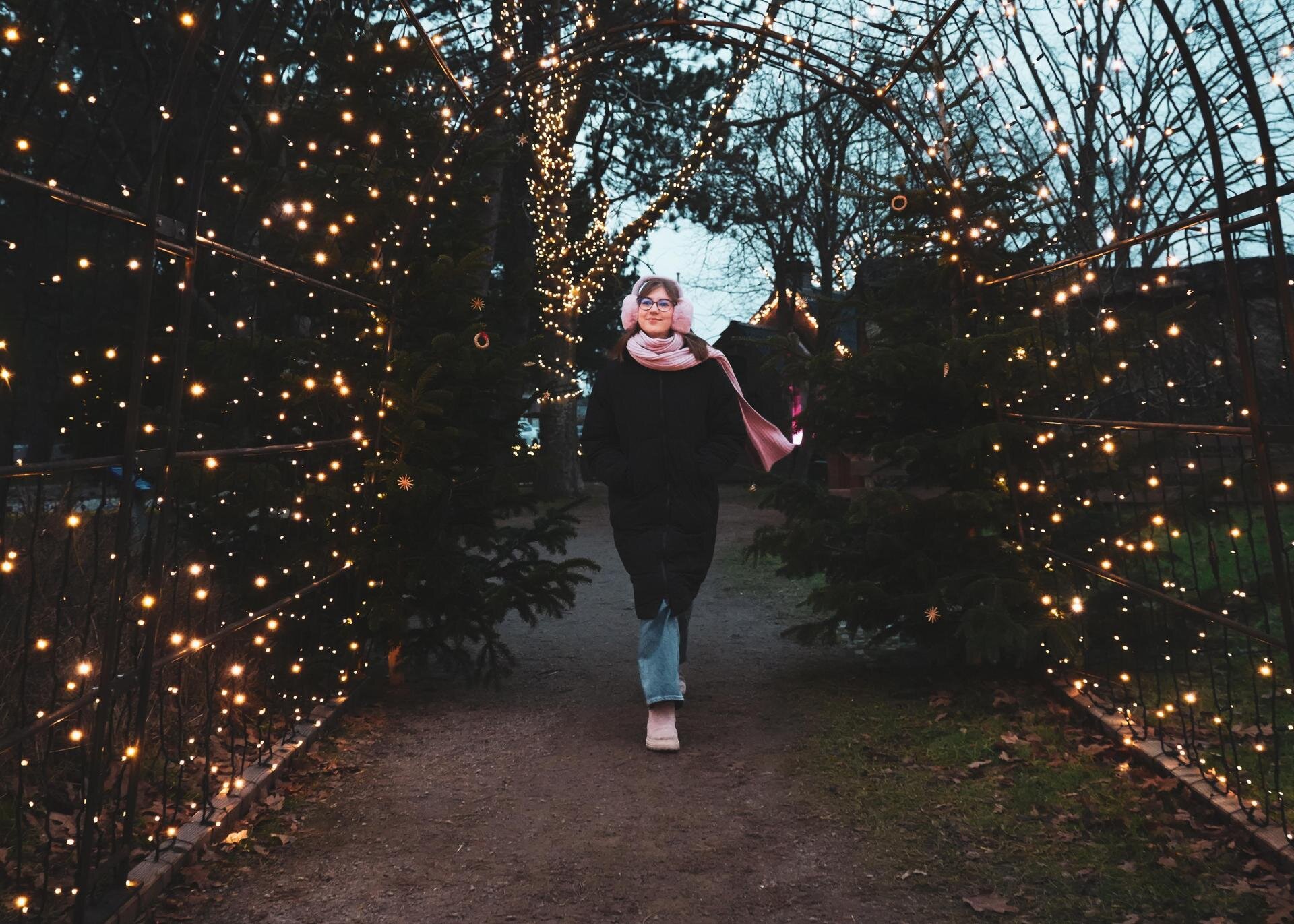 Eine junge Frau, eingepackt in Mütze und Schal, schlendert durch einen Lichtertunnel zum Weihnachtsmarkt.