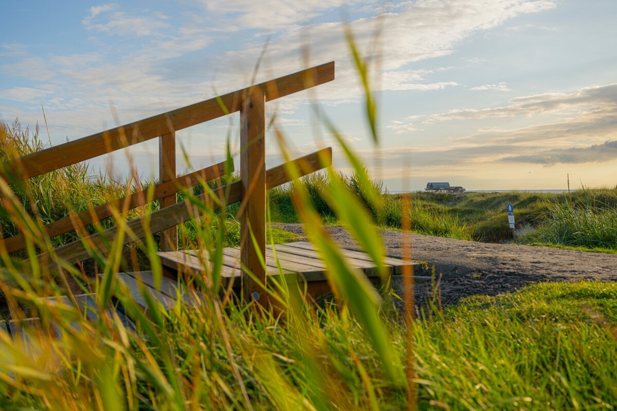 Blick über den Deich durch ein Treppengeländer in Richtung Pfahlbau am Strand